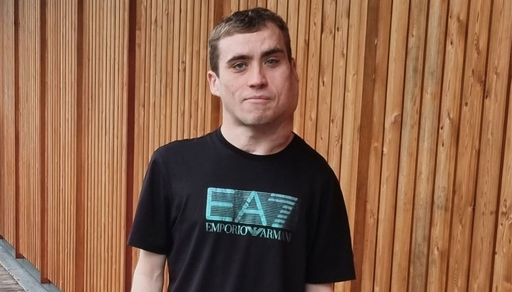 A young man in black T-shirt stands in front of an exterior wall made from wooden cladding.