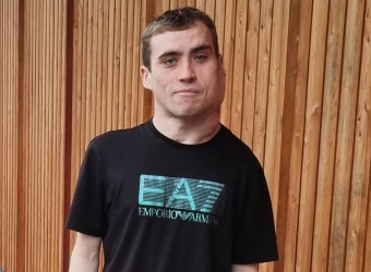 A young man in black T-shirt stands in front of an exterior wall made from wooden cladding.