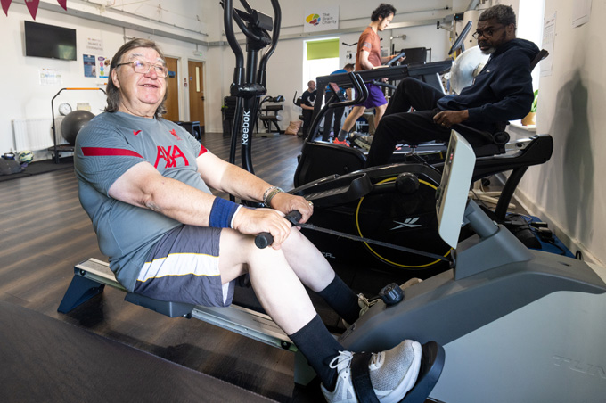 Men exercising in gym. A man on a rowing machine is in the foreground. He looks happy