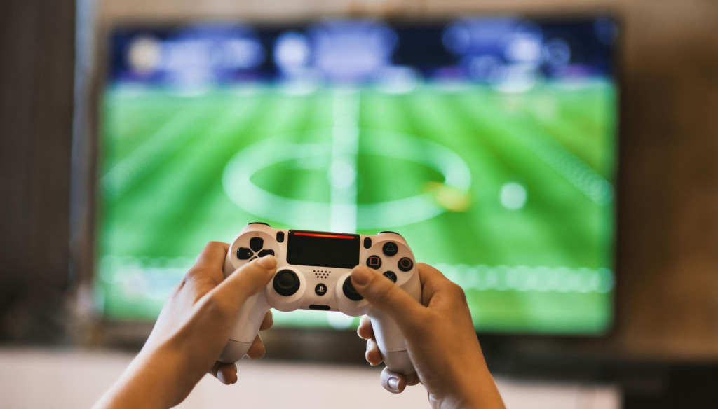 Hands holding a games controller with a football game on the TV in the background