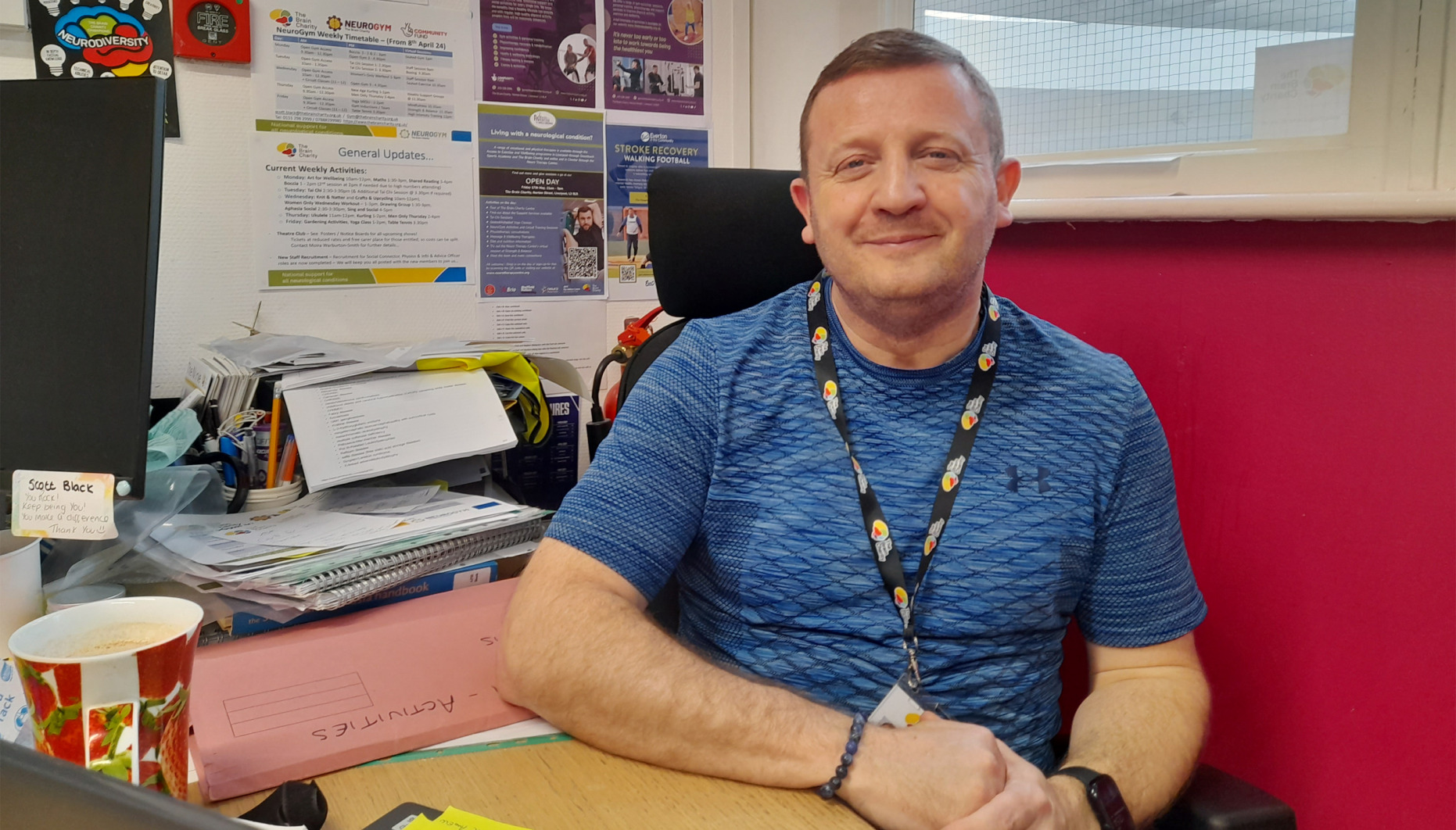 Man sitting behind a desk wearing a Brain Charity lanyard. He is smiling at the camera.