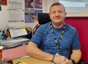 Man sitting behind a desk wearing a Brain Charity lanyard. He is smiling at the camera.