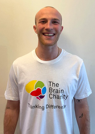 A man, Archie, in a Brain Charity T shirt standing in front of a white wall, smiling