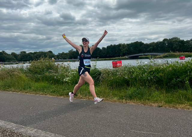 A woman in running gear, cap and sunglasses does a star jump as she runs along a riverside path