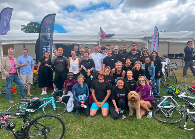 A group of people are gathered in front of a white tent with event flags/banners. Many participants are wearing athletic gear. Several bicycles are laid out in front and to the side of of the group. A fluffy dog sits with the group in the front row. The photo is taken outside on a partly cloudy day with banner flags visible in the background