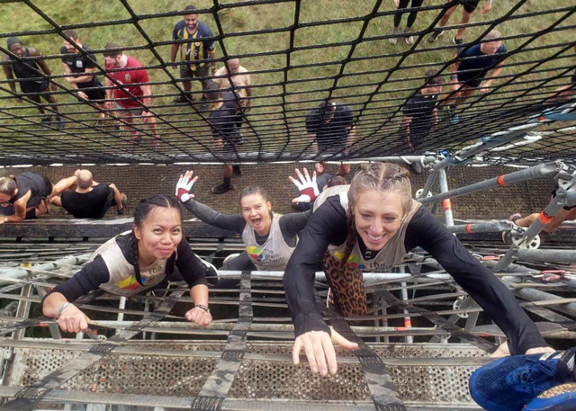 The Tough Mudder participants  wearing athletic gear and Brain Charity shirts, with expressions of effort and enjoyment as they navigate the challenging structure. The perspective creates an interesting geometric pattern of metal bars and supports.