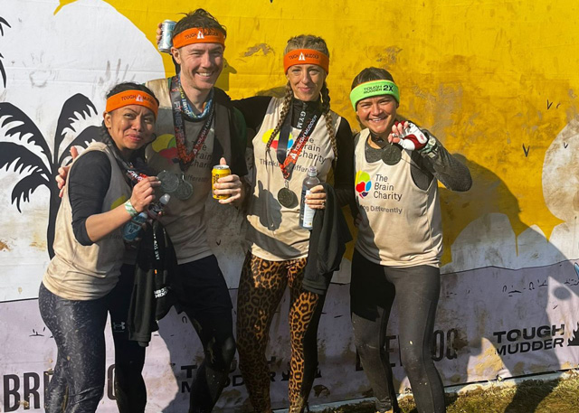 A man and three women celebrating at the end of the Tough Mudder challenge. They have medals, are wearing headbands and Brain Charity running shirts. They are very muddy