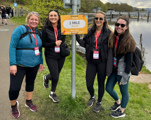 Four women in athletic wear stand next to a '1 MILE' distance marker sign along a waterfront path. They are all wearing event lanyards/badges and smiling at the camera. 