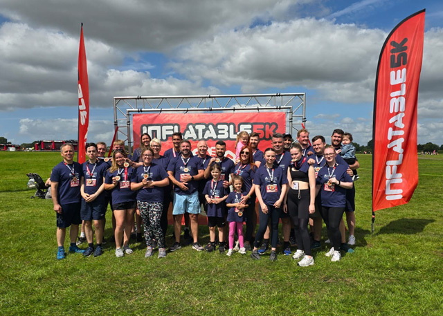 A group photo of participants after an 'Inflatable 5k' event, standing in front of a branded banner and red feather flags. The group is wearing athletic wear, with many displaying medals. They're gathered on a grassy field under a dramatic cloudy sky with patches of blue. 