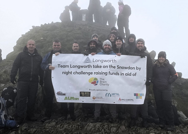 The group of staff from Longworth are dressed in mountain gear at the summit of Snowdon. They are holding a banner that says "Team Longworth take on the Snowdon by night challenge raising funds in aid of The Brain Charity