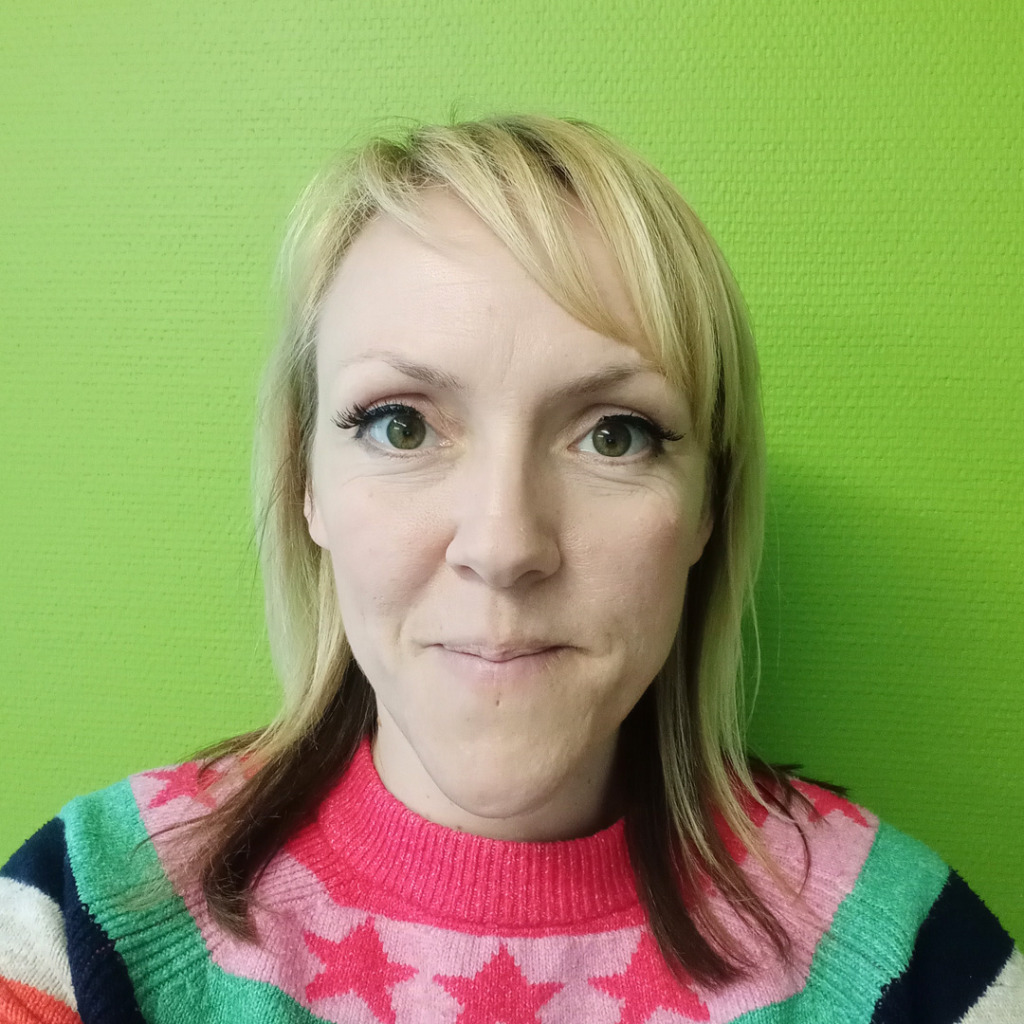 A woman with shoulder length hair in a colourful jumper against a plain green wall.