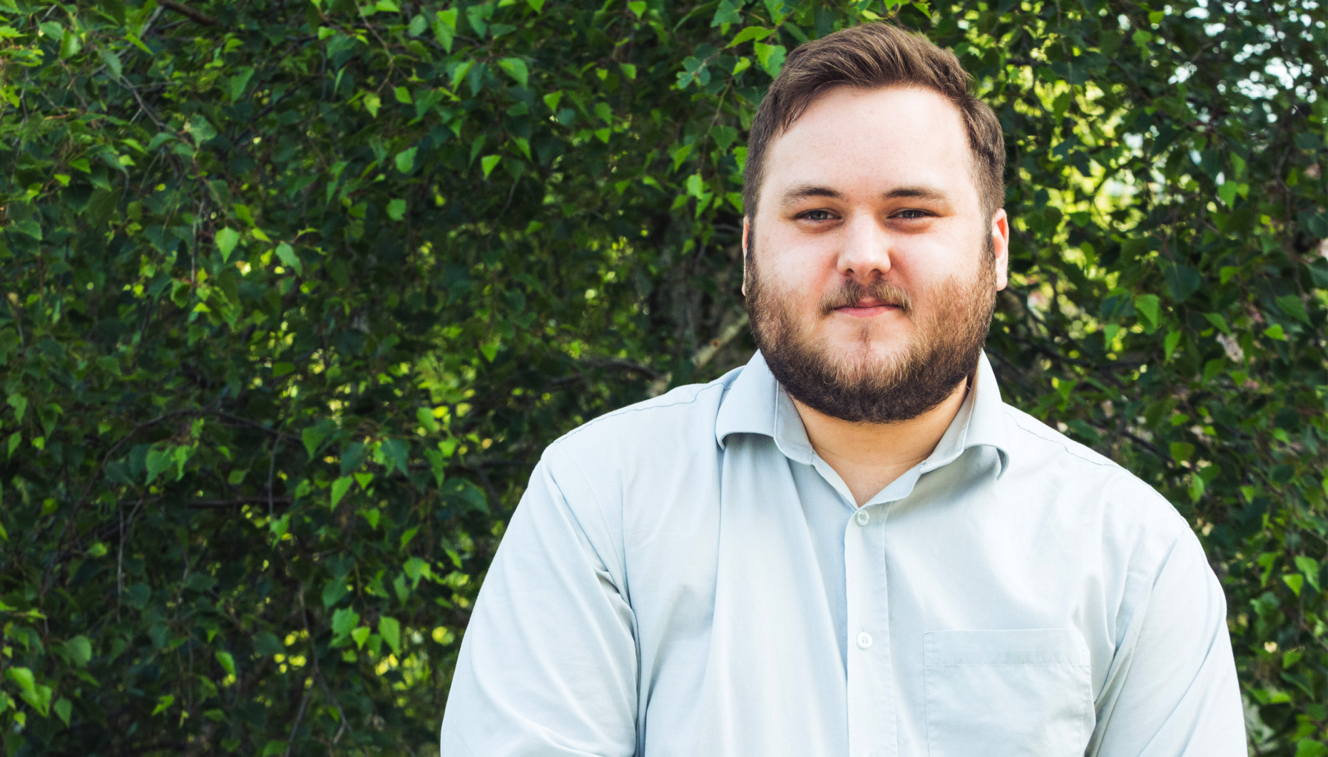 Young man with beard in an open necked shirt standing in front of a wall of Ivy