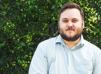 Young man with beard in an open necked shirt standing in front of a wall of Ivy