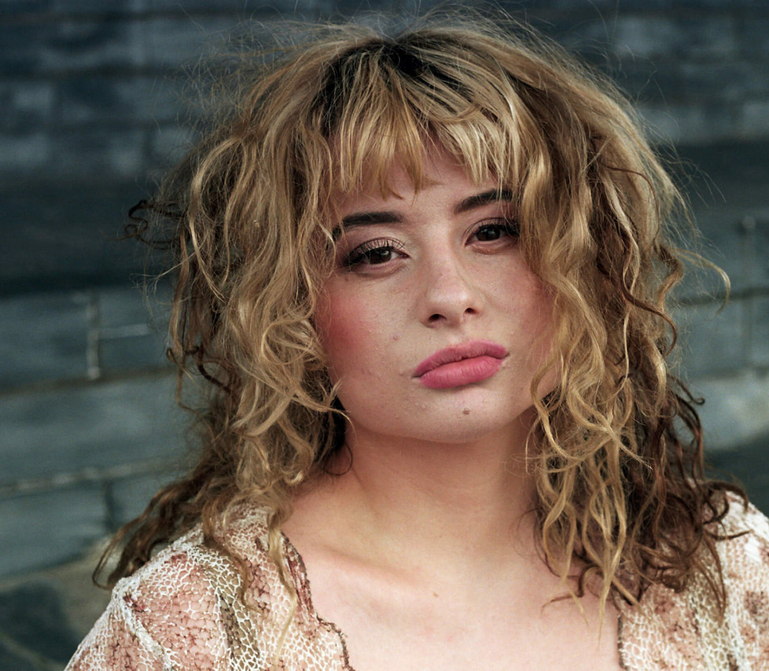 Woman with blond tousled hair standing against a bare stone wall