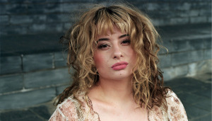 Woman with blond tousled hair standing against a bare stone wall