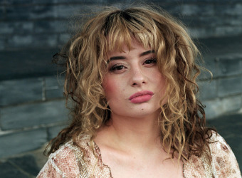 Woman with blond tousled hair standing against a bare stone wall