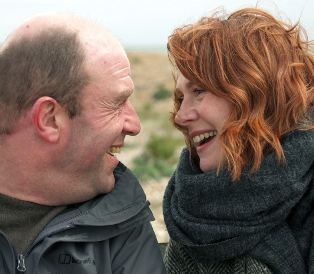 A man and a woman sitting in the countryside. They are looking at each other and smiling