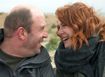 A man and a woman sitting in the countryside. They are looking at each other and smiling