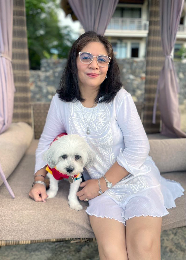 Woman with dark hair and glasses in a white dress, sitting on a couch with a small white dog