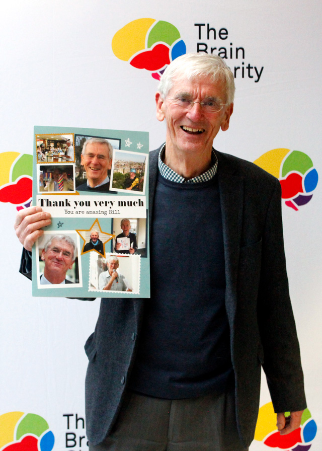 A man holding a large leaving card and smiling at the camera they are standing in front of a logo board with The Brain Charity logo