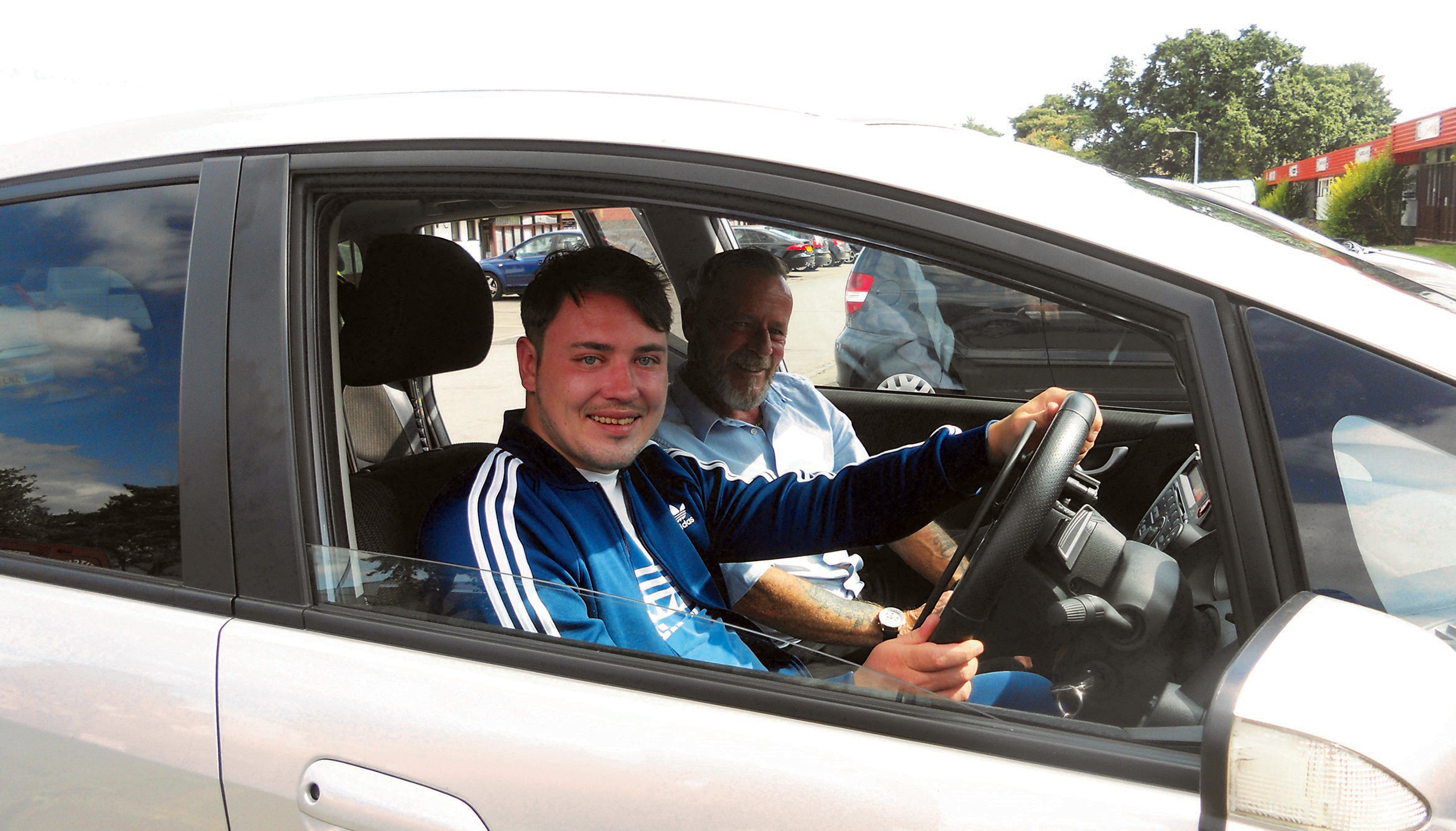 2 men sitting in the front seats of a car. The man in the driving seat is younger and they are both looking at the camera and smiling.