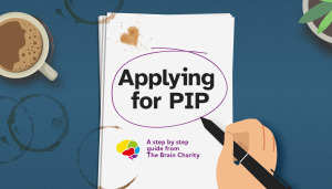 A notepad with the words Applying for PIP. A cartoon hand holding a pen has circled the title. There are coffee stains on it indicating all the hard work that has been put in to the application.