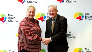 A woman and a man shaking hands and smiling at the camera they are standing in front of a logo board with The Brain Charity logo