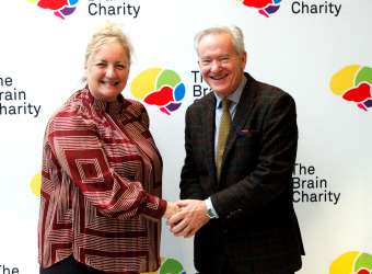 A woman and a man shaking hands and smiling at the camera they are standing in front of a logo board with The Brain Charity logo