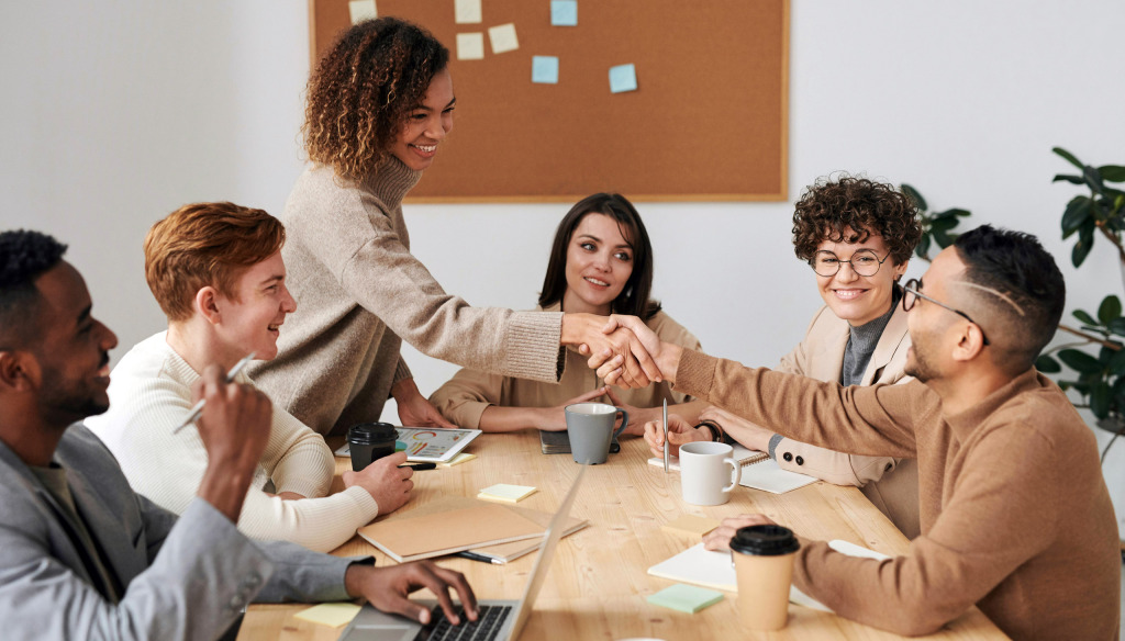 People sitting around a table in a work meeting. One woman is standing up and shaking hands across the table with a seated male colleague