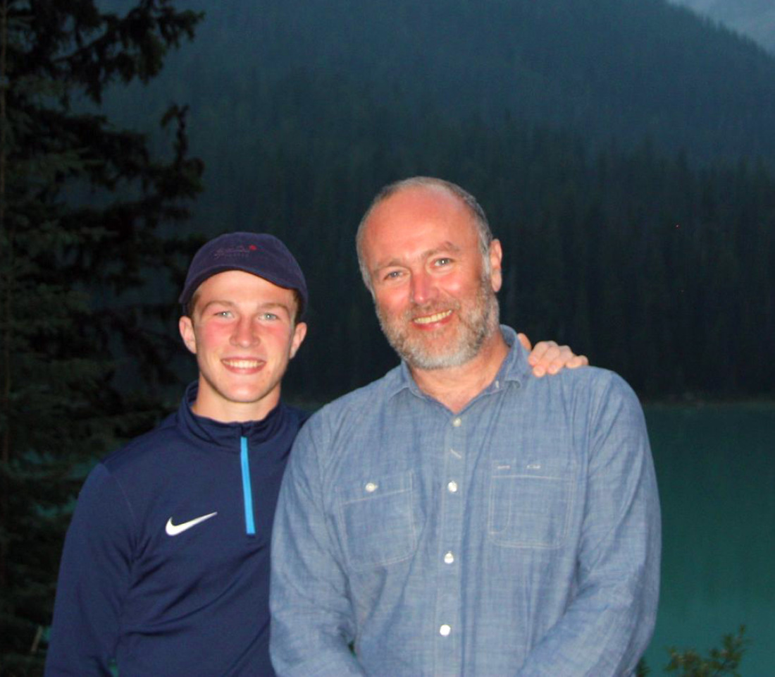 Archie with his dad Dave standing in front of a fjord at dusk. Archie has his left arm around his dads shoulder and they are both smiling at the camera