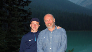 Archie with his dad Dave standing in front of a fjord at dusk. Archie has his left arm around his dads shoulder and they are both smiling at the camera