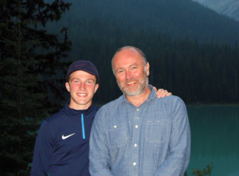 Archie with his dad Dave standing in front of a fjord at dusk. Archie has his left arm around his dads shoulder and they are both smiling at the camera