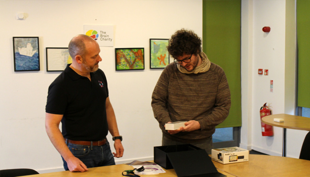 Chris, a young man stands looking at a piece of adaptive technology. A man in a black T shirt stands on the left explaining it.