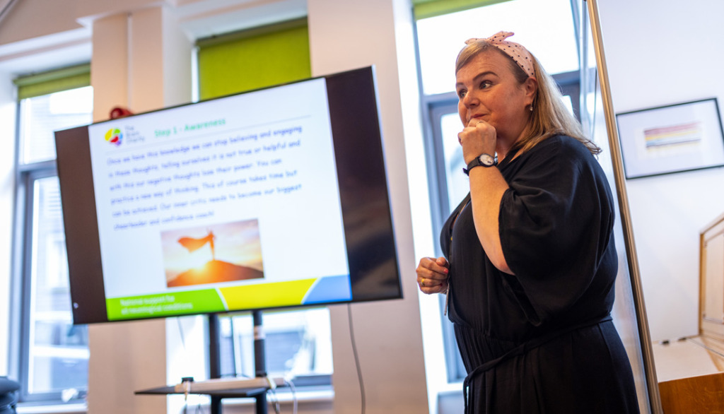 A trainer stands to deliver a course. To her left is a TV with a PowerPoint presentation on it.