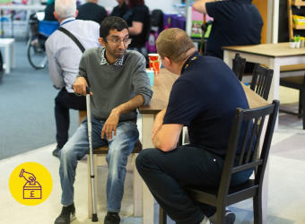 A seated man, Rupak, wearing grey top, jeans and glasses. He is sitting speaking to a support worker at The Brain Charity